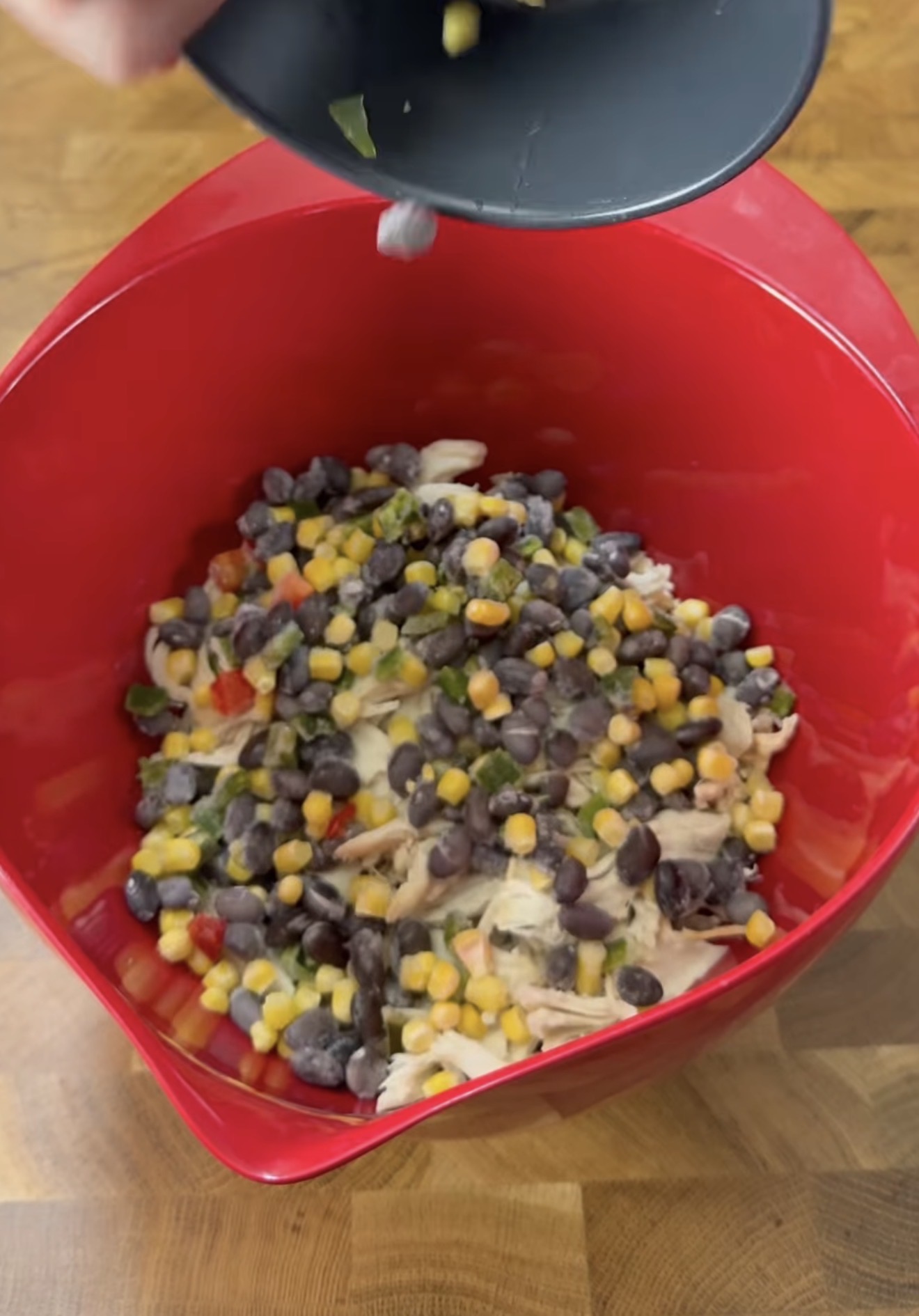 A red mixing bowl filled with black beans, corn, peppers, and shredded chicken in a creamy mixture. A hand is pouring more ingredients from a small black plate into the bowl. The bowl sits on a wooden surface.