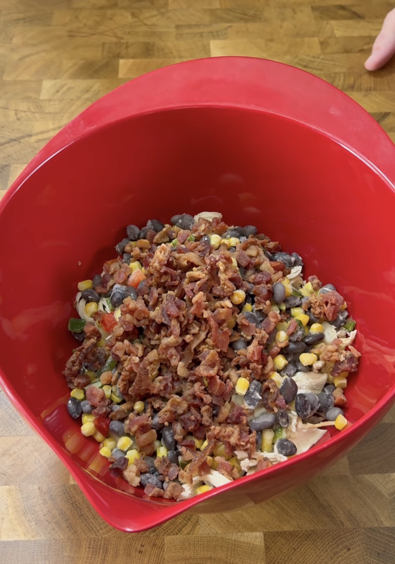 A red mixing bowl on a wooden counter contains a mixture of black beans, corn, chopped bacon, and other ingredients. A hand is visible in the background.