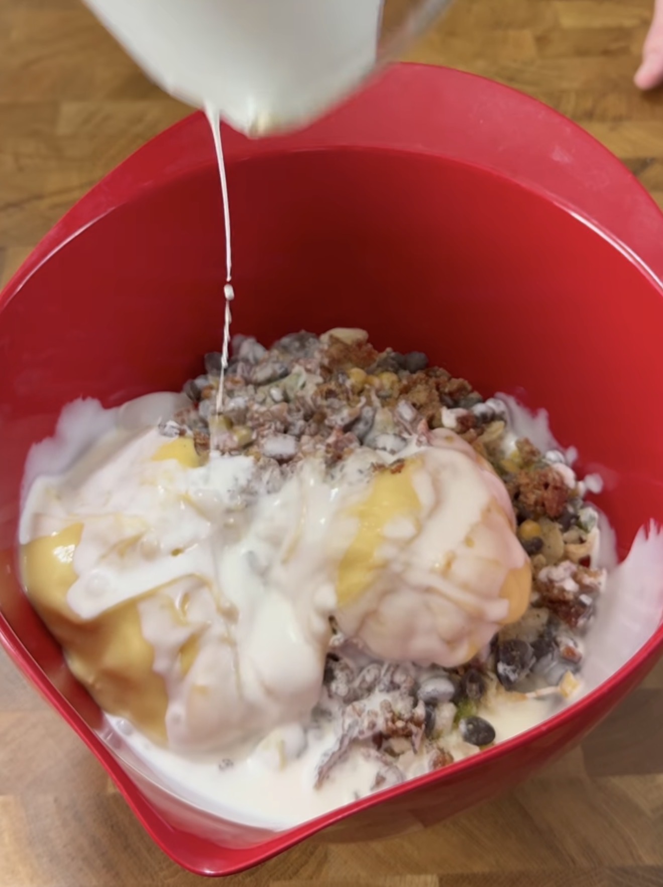 A red mixing bowl contains dried fruit, nuts, and dollops of yogurt, with milk being poured in from a jug above. The bowl is on a wooden surface.