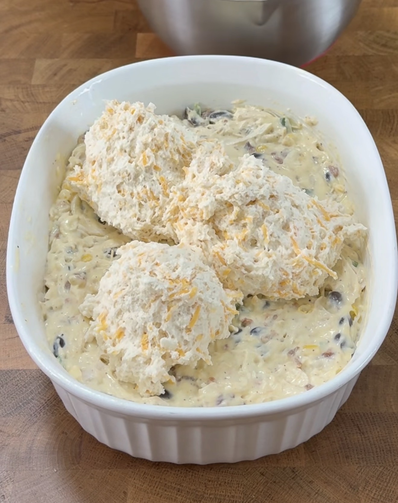 A white baking dish filled with a thick, chunky batter or casserole mixture, topped with three large scoops of biscuit-like dough, resting on a wooden countertop.