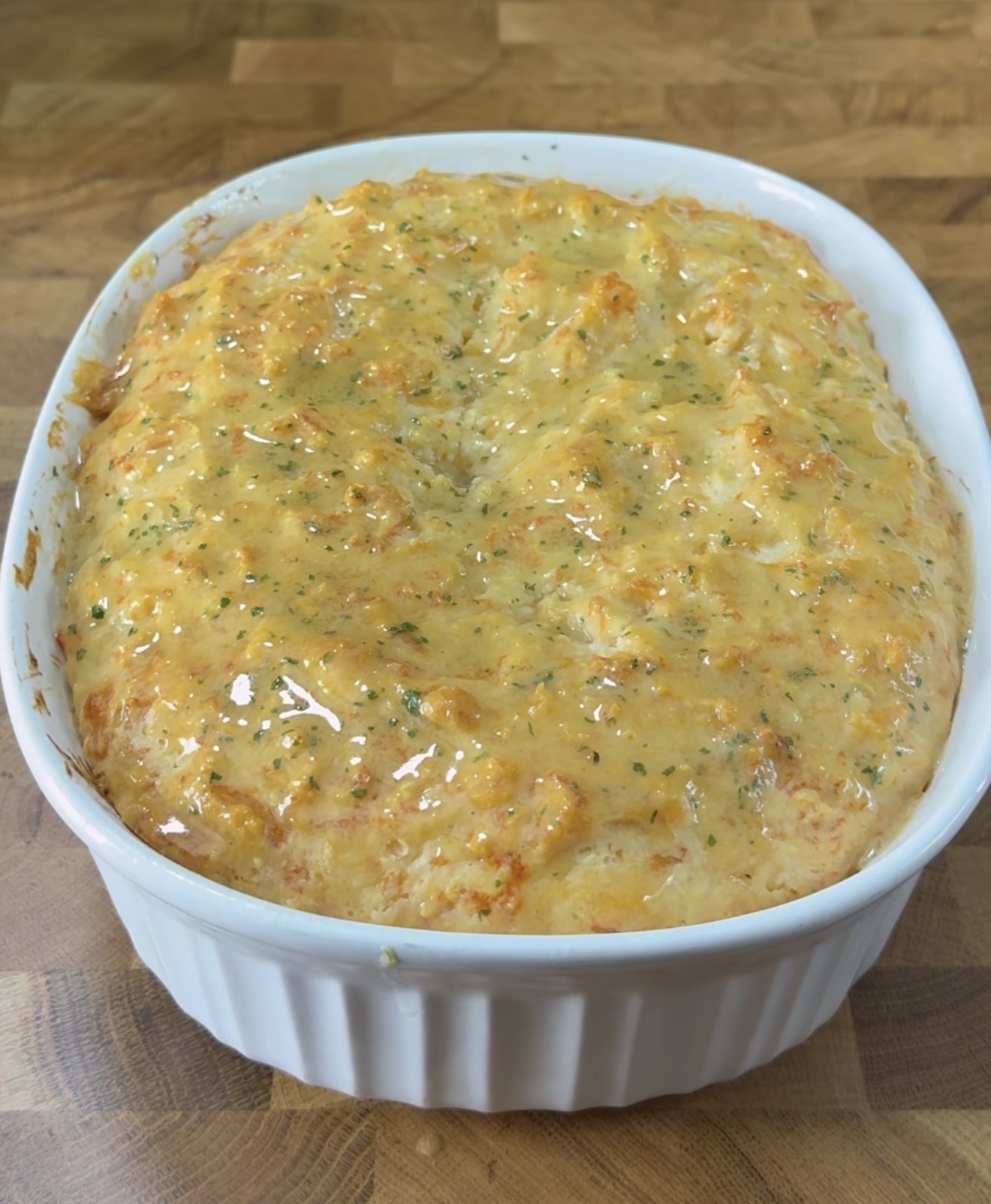 A golden-brown, cheesy baked casserole sits in a white oval dish on a wooden surface, with a glossy, herb-speckled top.