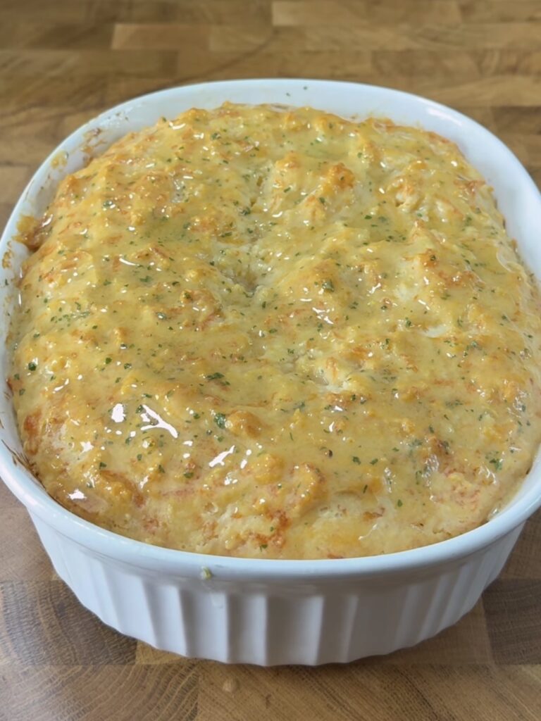Golden, cheesy bread baked in a white ceramic dish, with a bubbly, slightly browned top and flecks of green herbs, resting on a wooden surface.