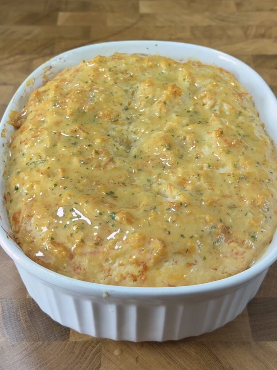 Golden, cheesy bread baked in a white ceramic dish, with a bubbly, slightly browned top and flecks of green herbs, resting on a wooden surface.