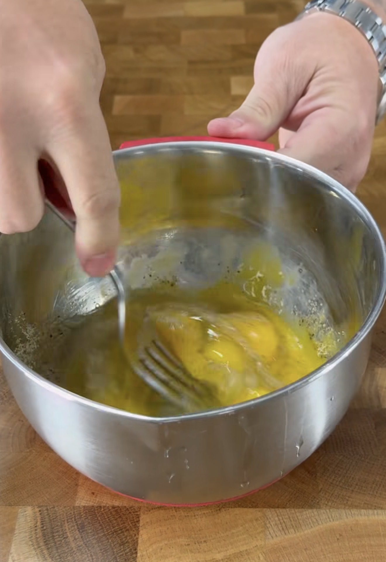 A person is whisking eggs with a fork in a stainless steel bowl on a wooden countertop.