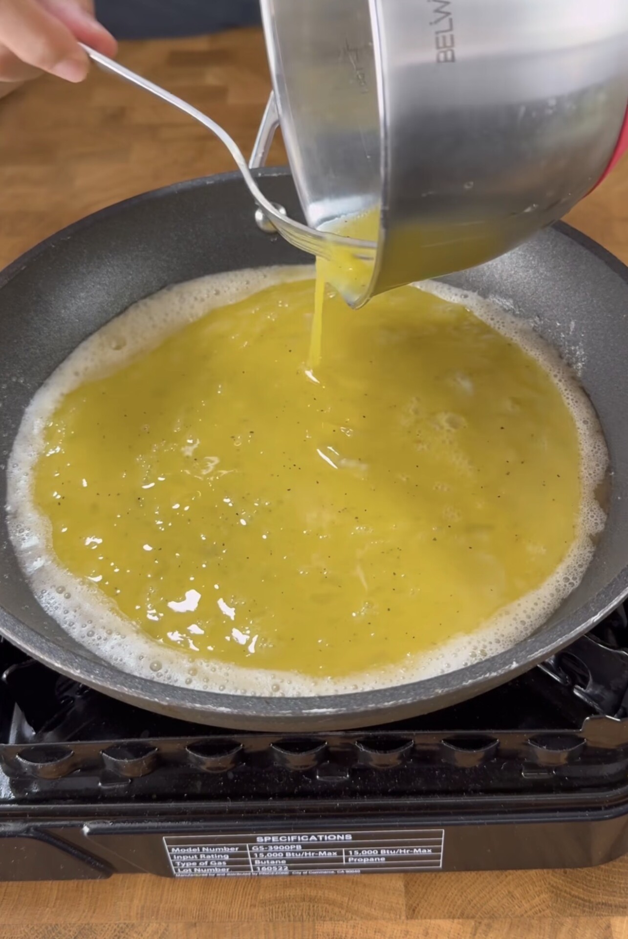 A hand pours beaten eggs from a metal bowl into a nonstick pan on a stovetop, preparing to cook scrambled eggs.