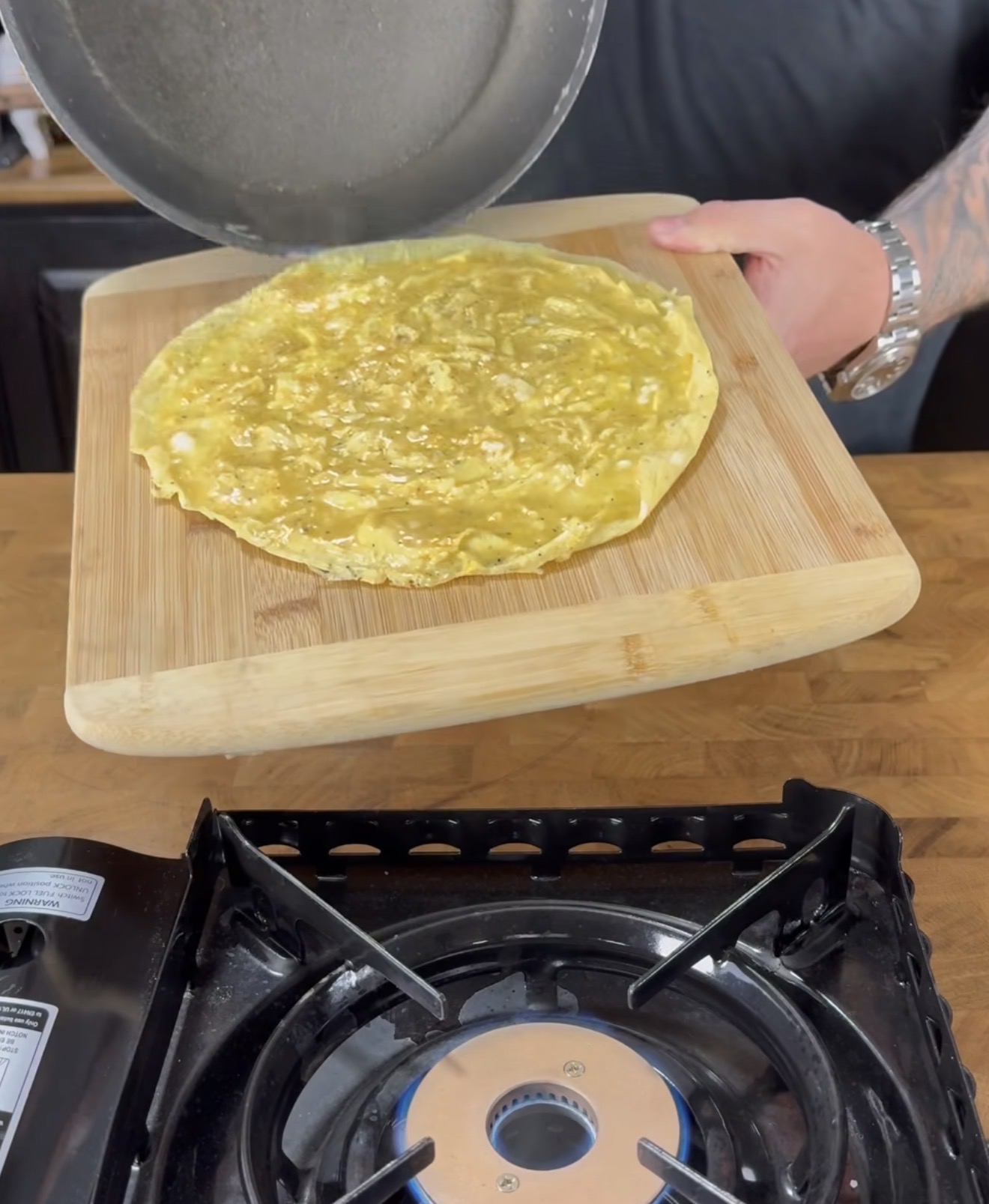 A person slides a cooked omelet from a pan onto a wooden cutting board above a lit gas stove.