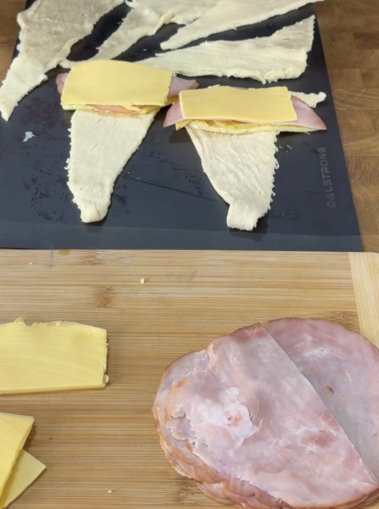 Two uncooked crescent roll dough triangles on a black cutting board are topped with slices of ham and cheese. More cheese slices and ham are on a wooden board below.