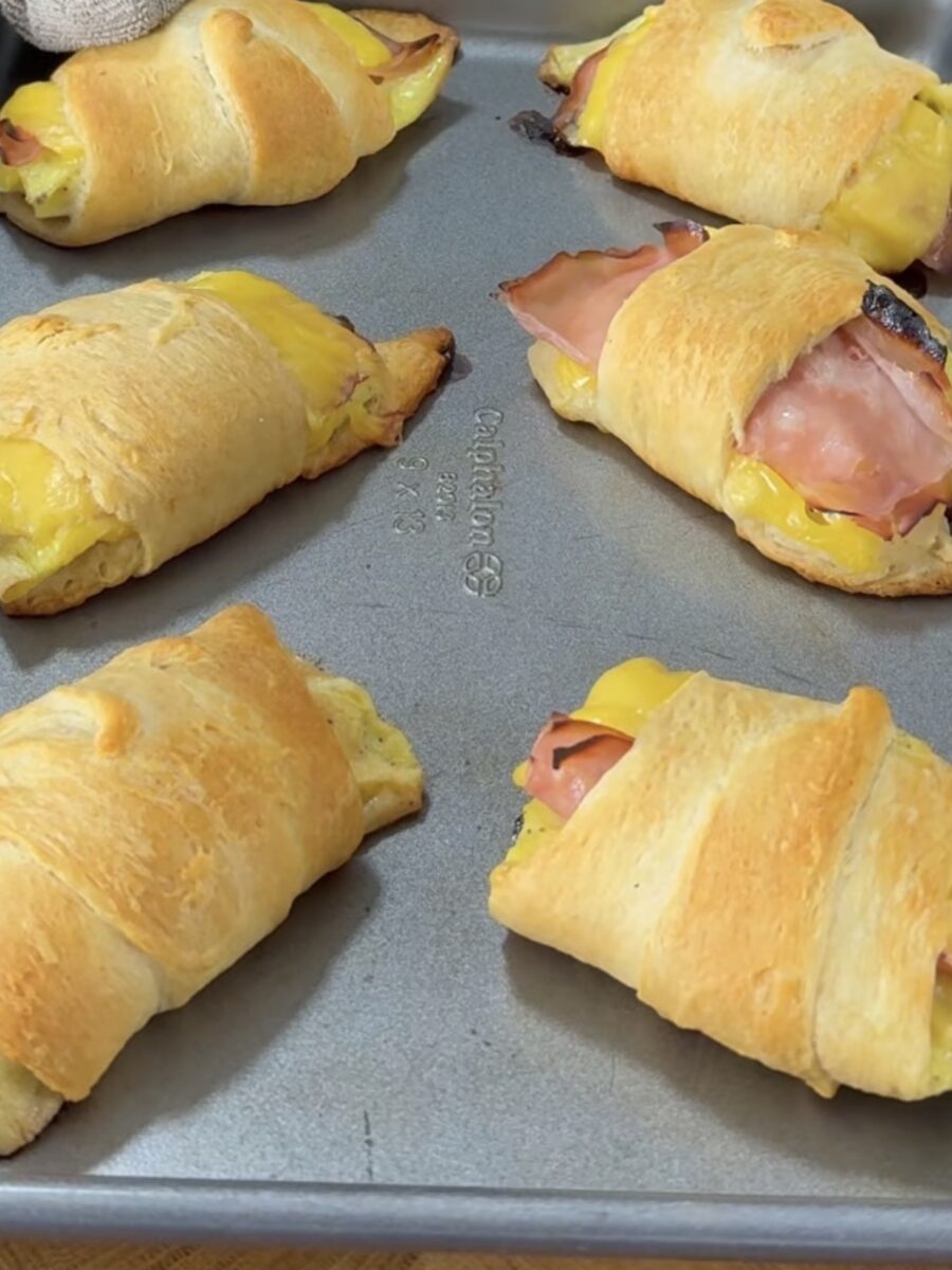 Six golden-brown crescent rolls filled with ham and melted cheese are arranged on a baking sheet. Some filling is slightly visible from the ends of the rolls.