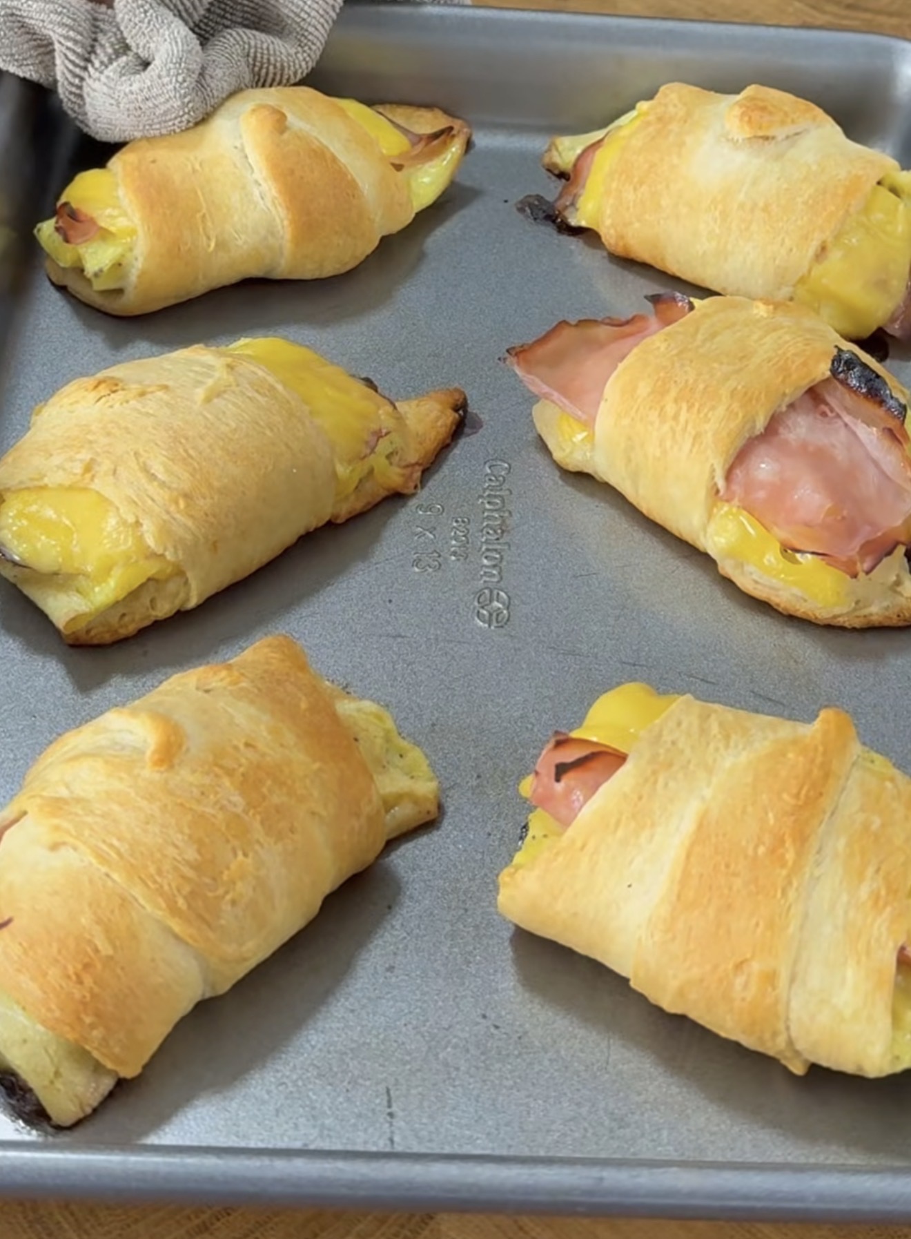 Six golden-brown crescent rolls filled with ham and melted cheese are arranged on a baking tray. One roll shows cheese oozing out, and a towel is visible in the corner.
