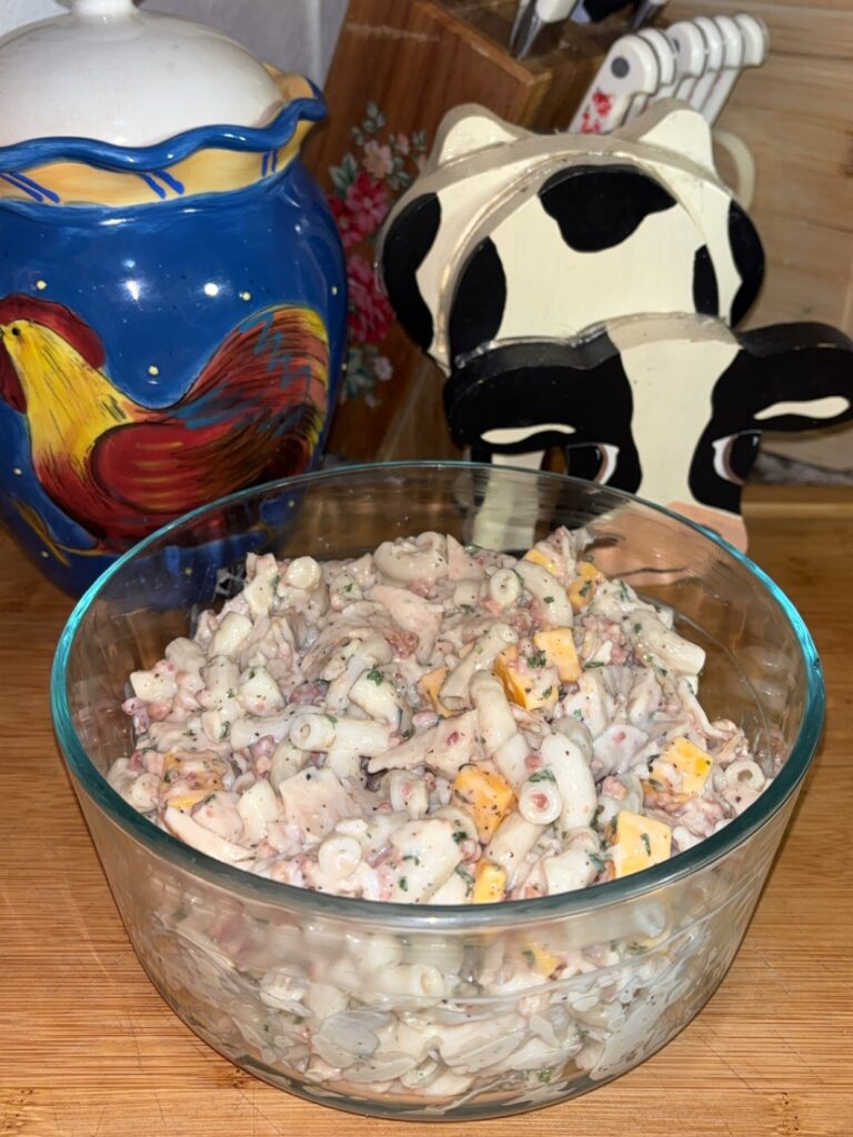A glass bowl filled with creamy pasta salad containing diced cheese sits on a wooden countertop, with a blue rooster canister and a cow-shaped napkin holder in the background.
