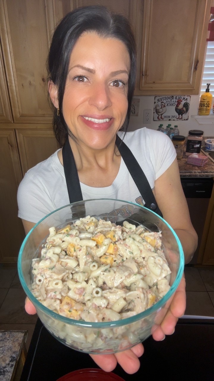A woman with dark hair smiles and holds a large glass bowl filled with creamy macaroni salad in a kitchen setting.