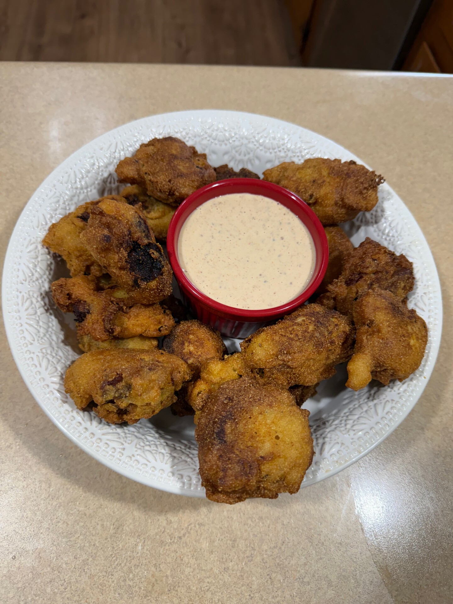 A white bowl filled with golden-brown, crispy fritters arranged around a small red ramekin of creamy dipping sauce, placed on a light-colored countertop.