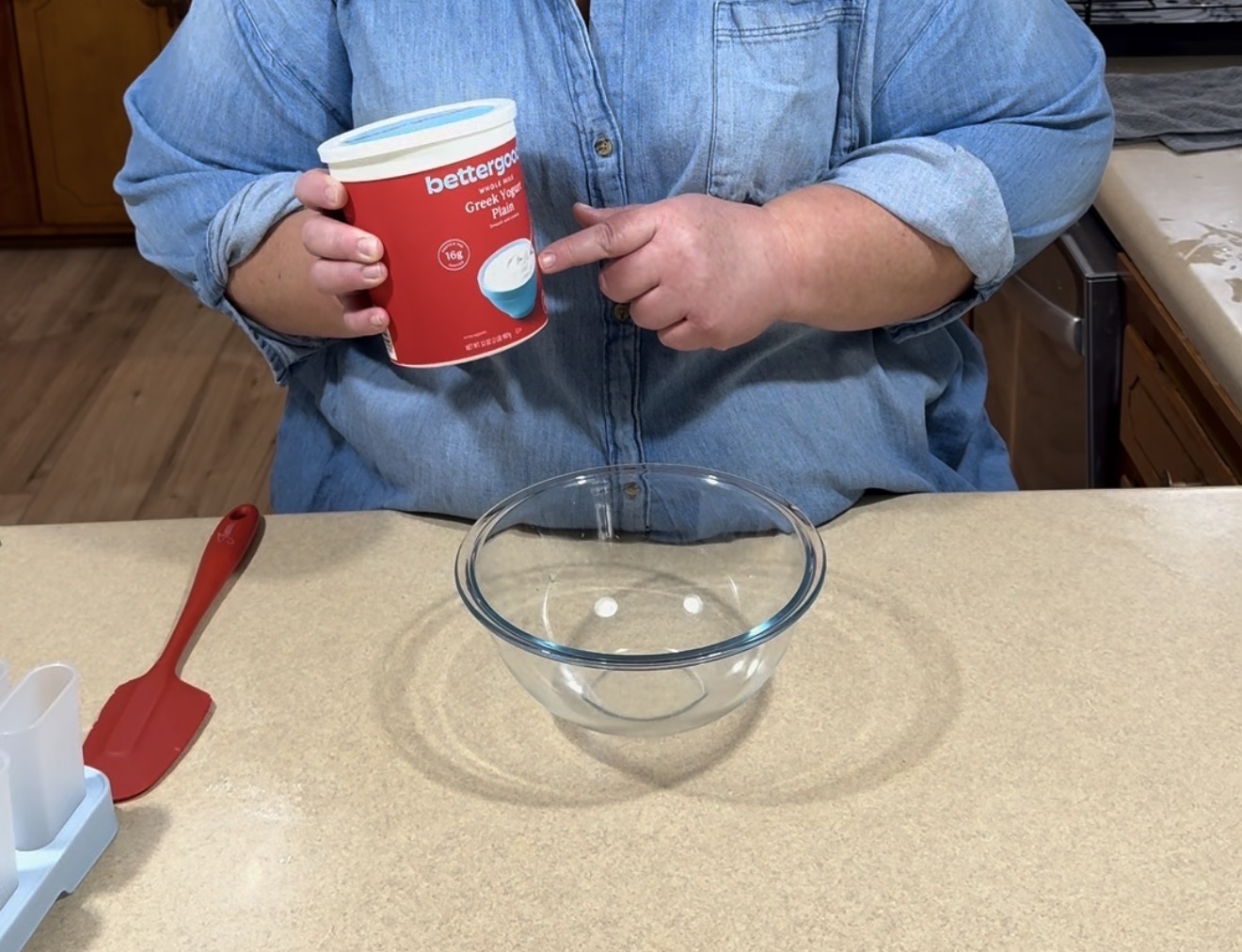 A person wearing a denim shirt holds a container of Greek yogurt above a clear glass mixing bowl on a kitchen counter with a red spatula nearby.
