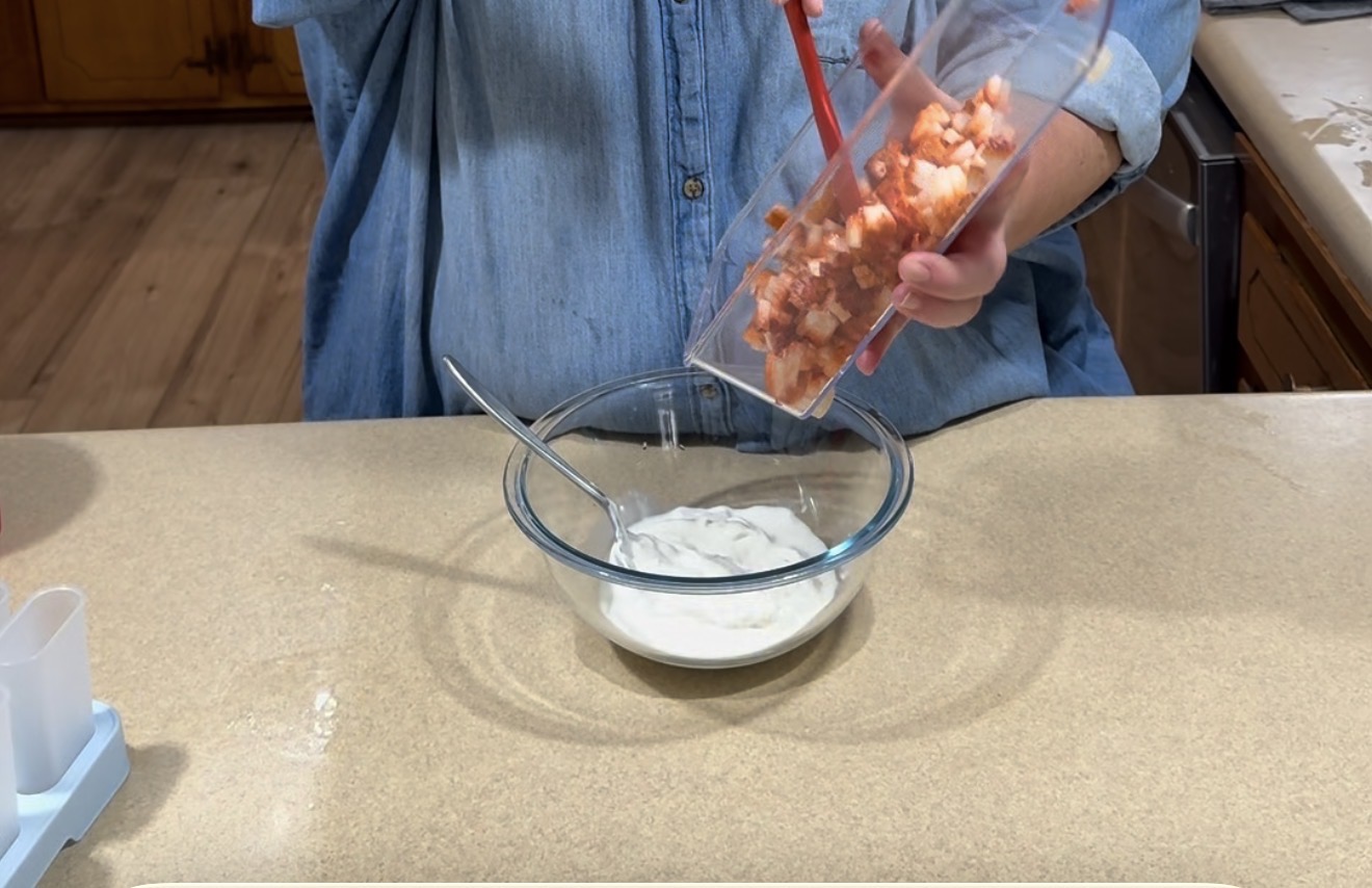 A person in a blue shirt pours chopped strawberries from a container into a glass bowl filled with yogurt on a kitchen counter. A spoon rests in the bowl.