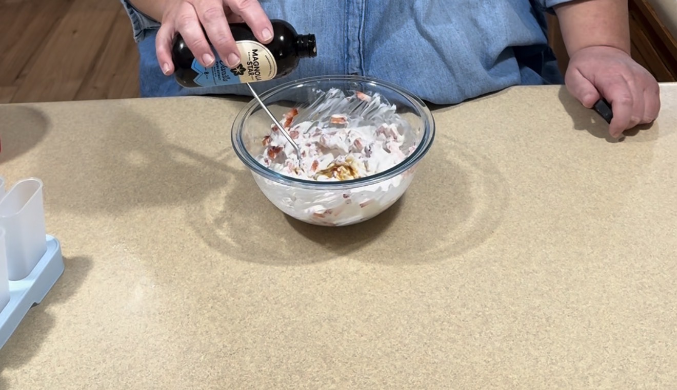 A person in a blue shirt is pouring Worcestershire sauce into a glass bowl containing a creamy mixture with chopped ingredients, on a beige countertop.