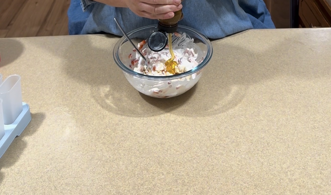 A person pours honey from a bottle into a glass bowl filled with a mixture that includes chopped ingredients and a creamy sauce, on a light-colored kitchen countertop.
