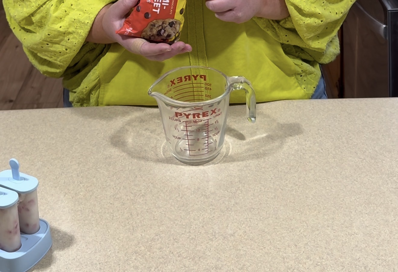 A person in a yellow shirt is holding a bag of mini chocolate chips over an empty glass measuring cup on a kitchen counter. A popsicle mold with popsicles is visible to the left.