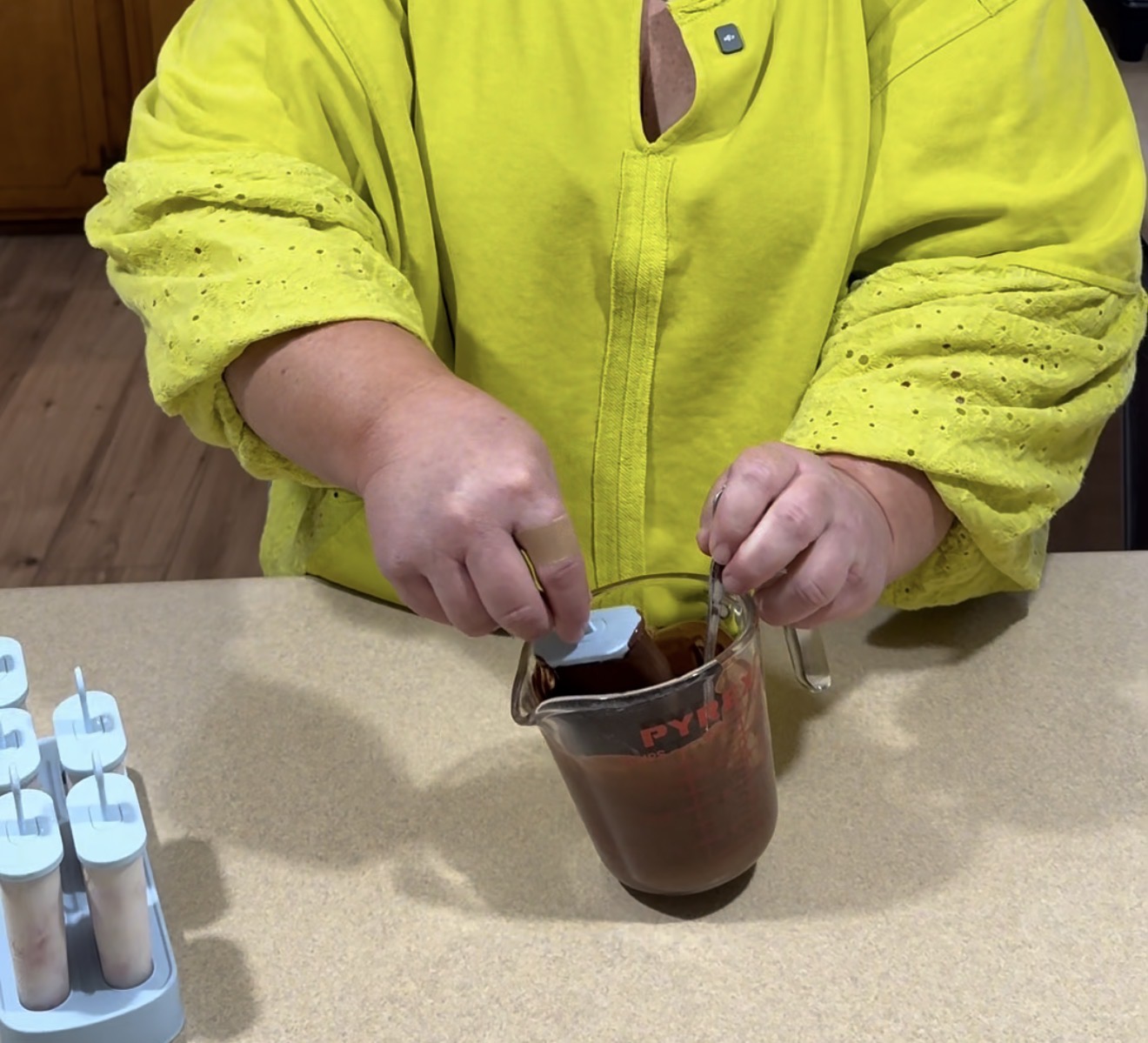 A person in a bright yellow shirt stirs a chocolate mixture in a glass measuring cup on a kitchen counter, with popsicle molds visible on the left.