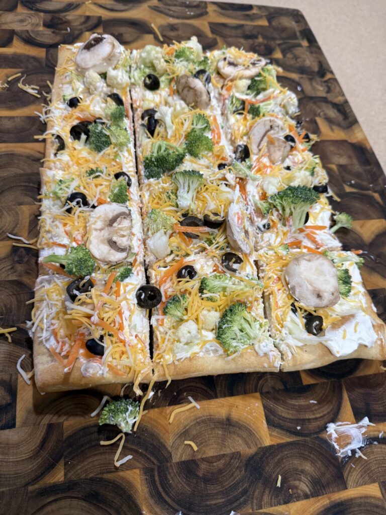 A rectangular vegetable pizza topped with shredded cheese, broccoli, mushrooms, black olives, carrots, and cauliflower, sliced into pieces on a patterned wooden cutting board.