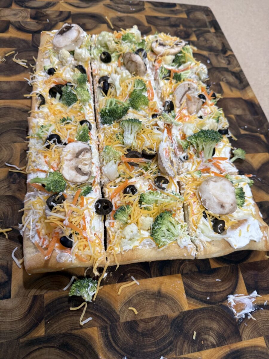 A rectangular vegetable pizza topped with shredded cheese, broccoli, mushrooms, black olives, carrots, and cauliflower, sliced into pieces on a patterned wooden cutting board.