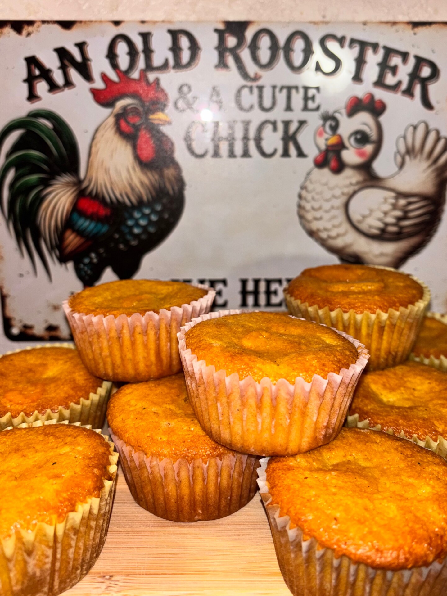 A group of golden-brown muffins is displayed on a wooden surface in front of a vintage sign featuring a colorful rooster and a white hen, with the text An Old Rooster & A Cute Chick Live Here.