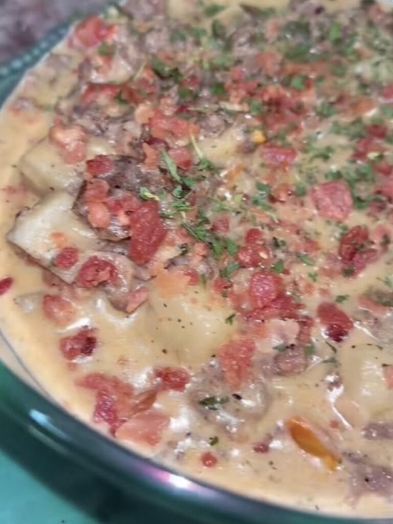 A close-up of a creamy casserole topped with diced tomatoes, ground meat, potatoes, herbs, and seasonings in a glass baking dish.