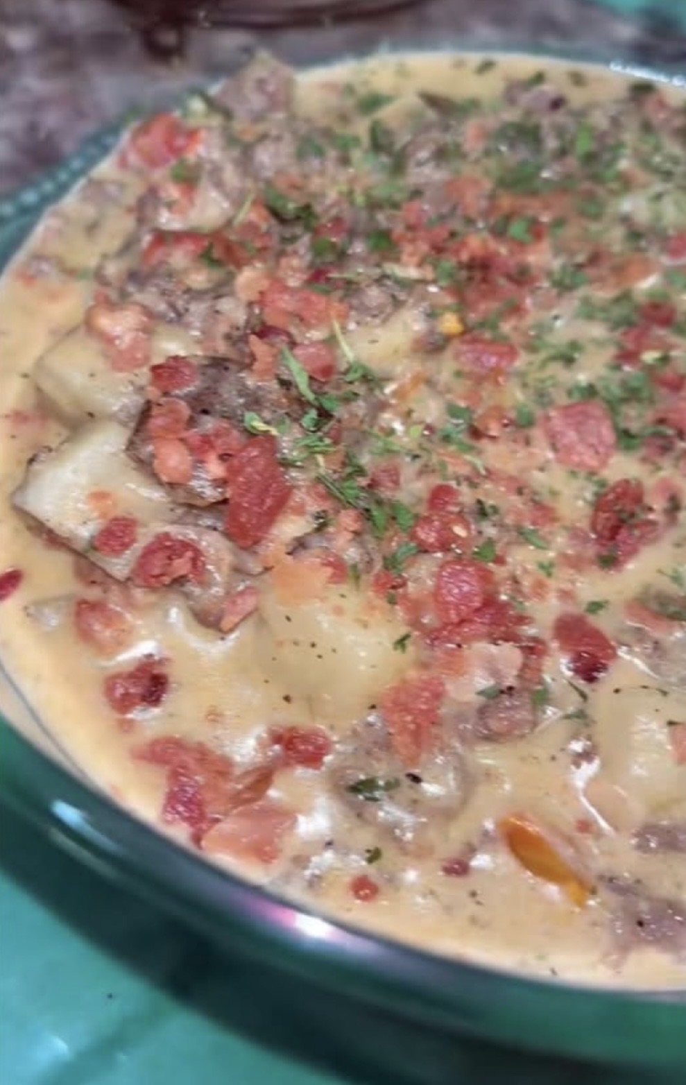A close-up of a creamy soup or stew with chunks of potato, ground meat, diced tomatoes, and herbs, served in a clear bowl. The dish is garnished with chopped greens.