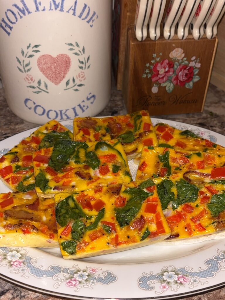 A plate of rectangular egg frittata slices with spinach, red bell pepper, and mushrooms sits on a floral-patterned plate. A decorative cookie jar and a wooden utensil holder are in the background.
