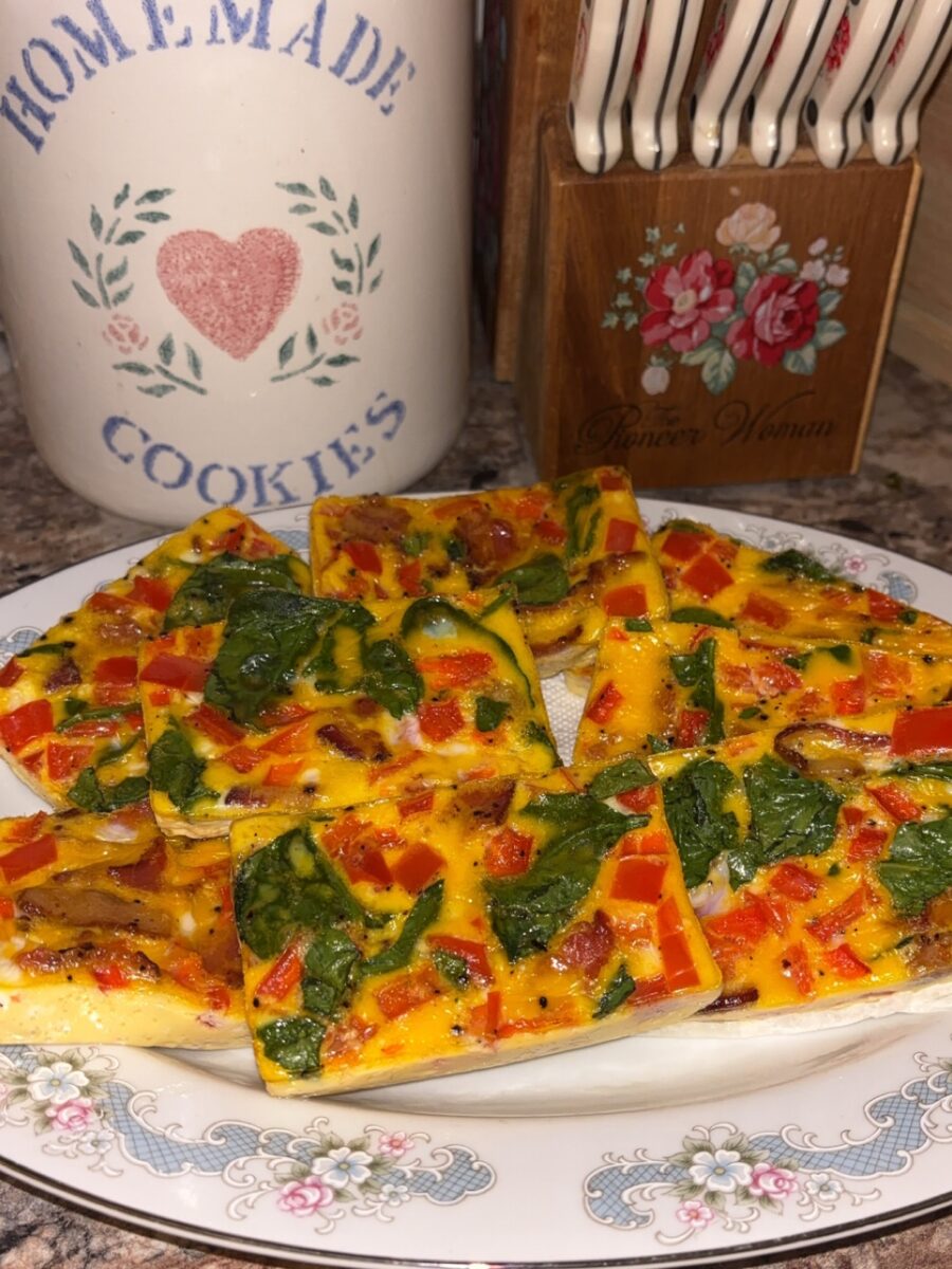 A plate of rectangular egg frittata slices with spinach, red bell pepper, and mushrooms sits on a floral-patterned plate. A decorative cookie jar and a wooden utensil holder are in the background.