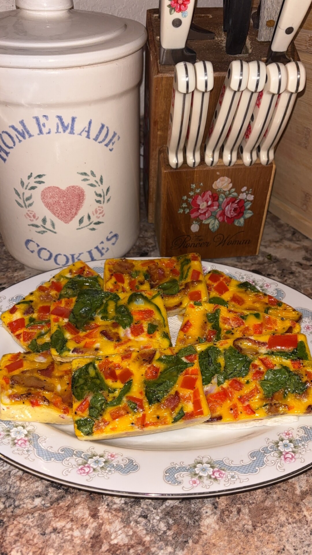 A plate of colorful baked egg squares with spinach, red peppers, and mushrooms sits on a kitchen counter near a cookie jar and a knife block.