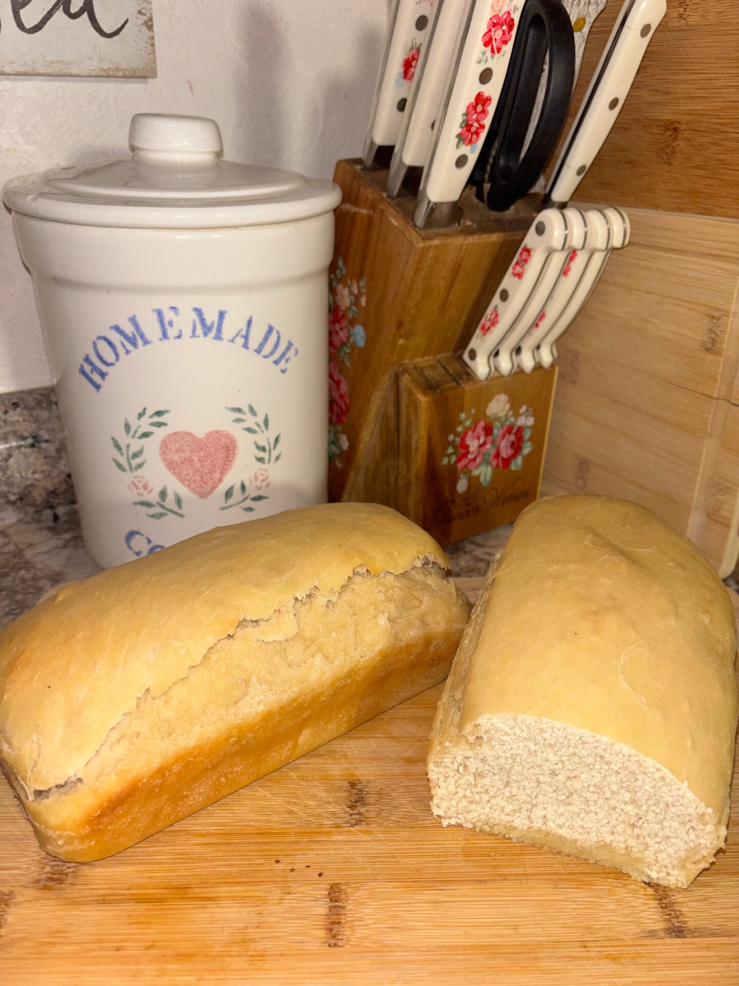 Two loaves of homemade bread sit on a wooden cutting board, one partially sliced. Behind them are a ceramic canister labeled Home Made, and two knife blocks with white-handled knives.