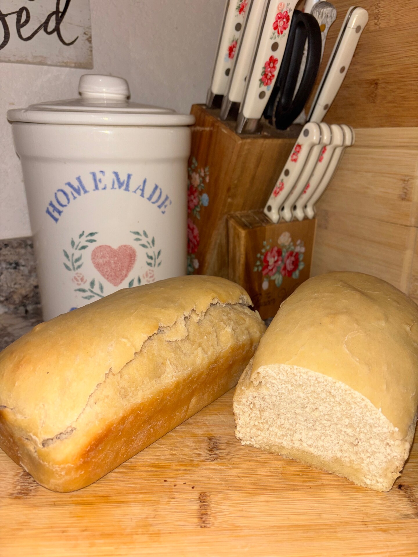 Two loaves of homemade bread, one sliced open to show the inside, sit on a wooden cutting board. Behind them are a utensil holder labeled homemade and a knife block with floral-patterned handles.