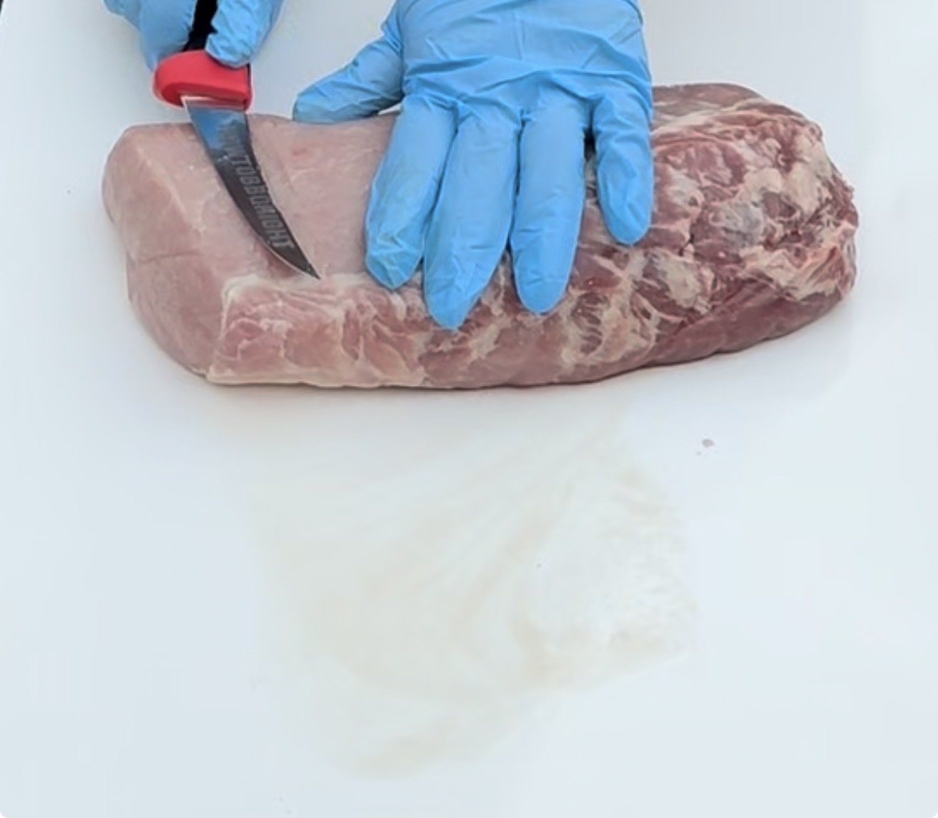 A person wearing blue gloves uses a knife to cut a large piece of raw pork on a white cutting board.