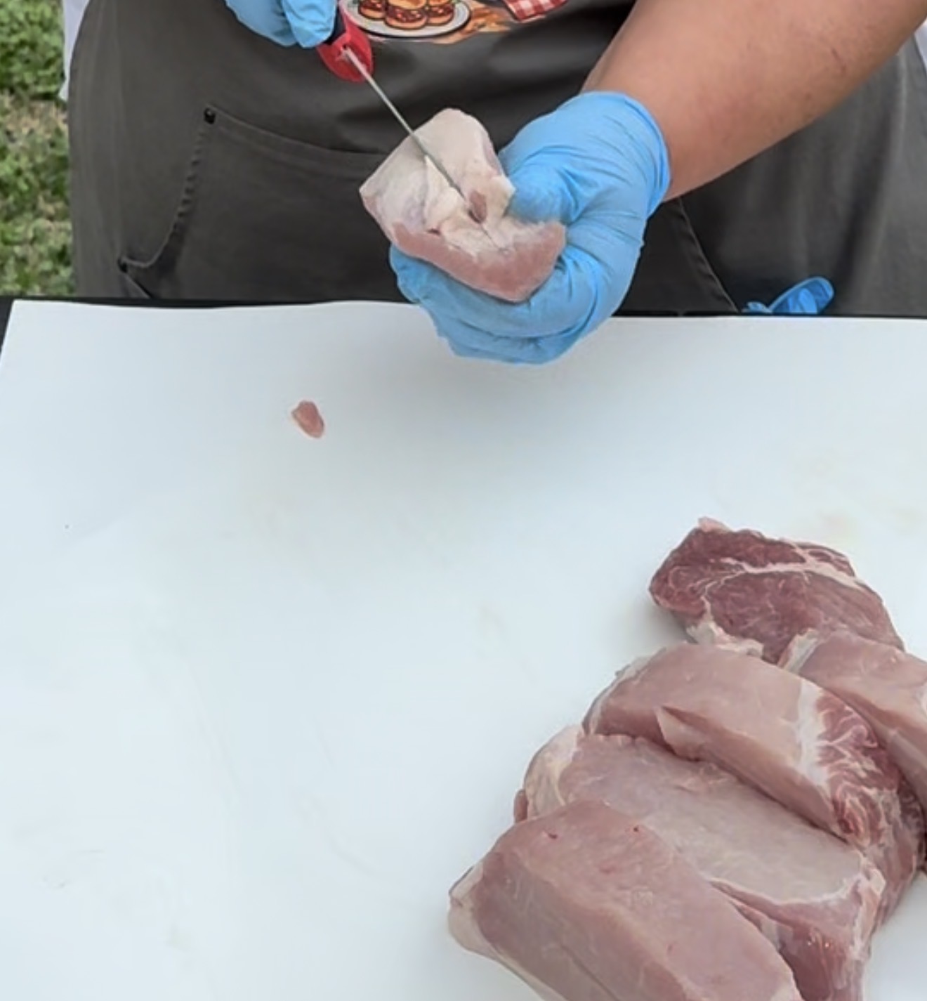 A person wearing blue gloves slices raw pork on a white cutting board outdoors, using a knife to cut the meat into thick pieces.