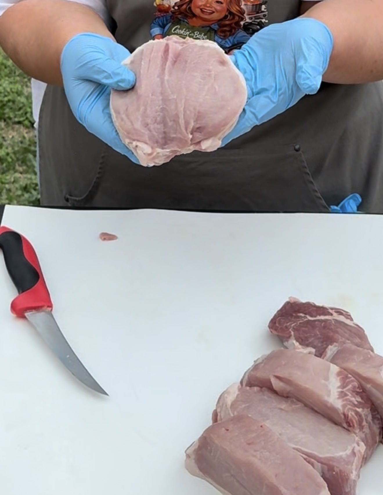 A person wearing blue gloves holds a raw pork chop above a white cutting board with a knife and several sliced pieces of pork on it.
