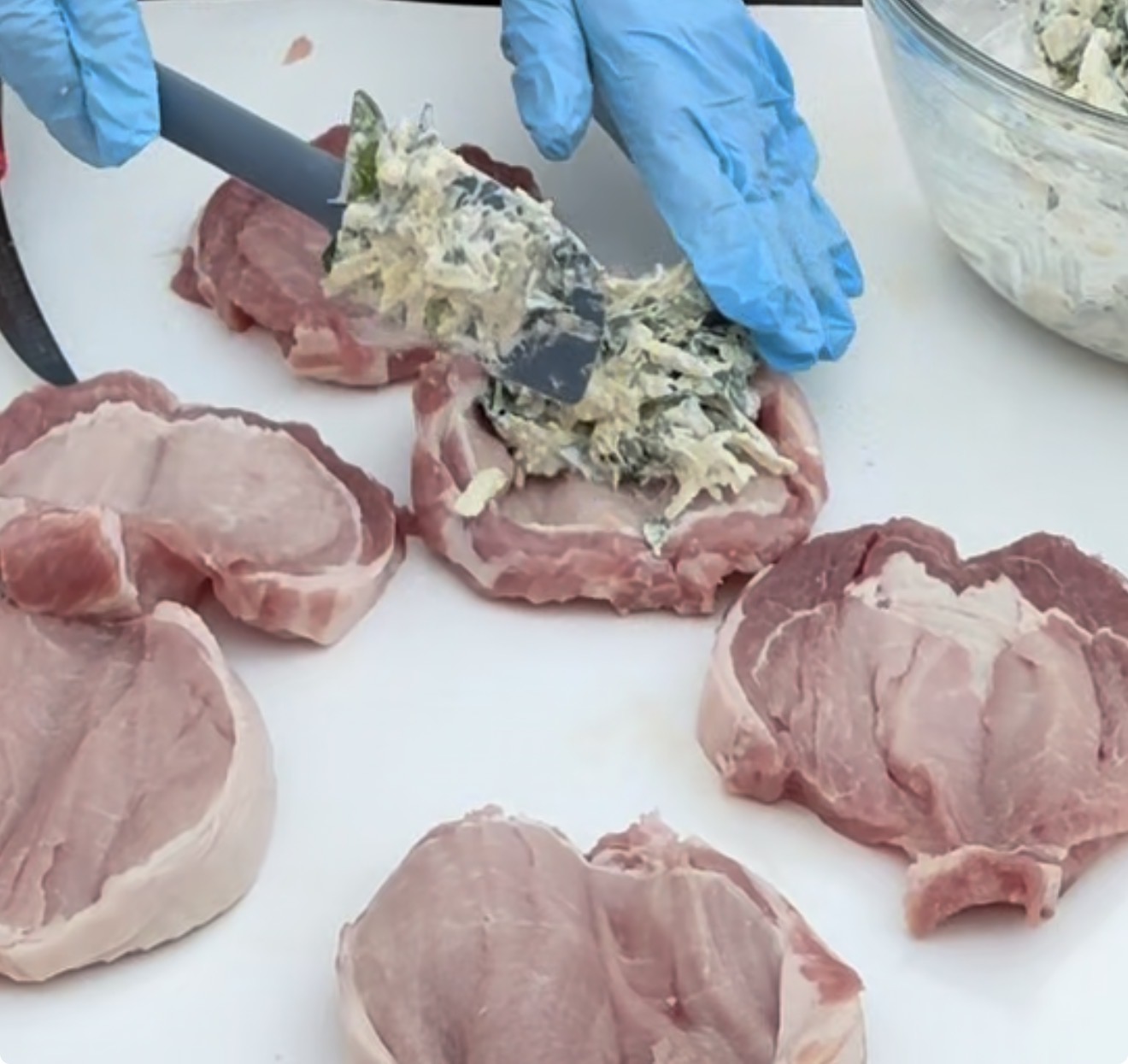 A person wearing blue gloves spreads a creamy mixture onto raw pork chops on a white cutting board, with more pork chops and a bowl of the mixture nearby.