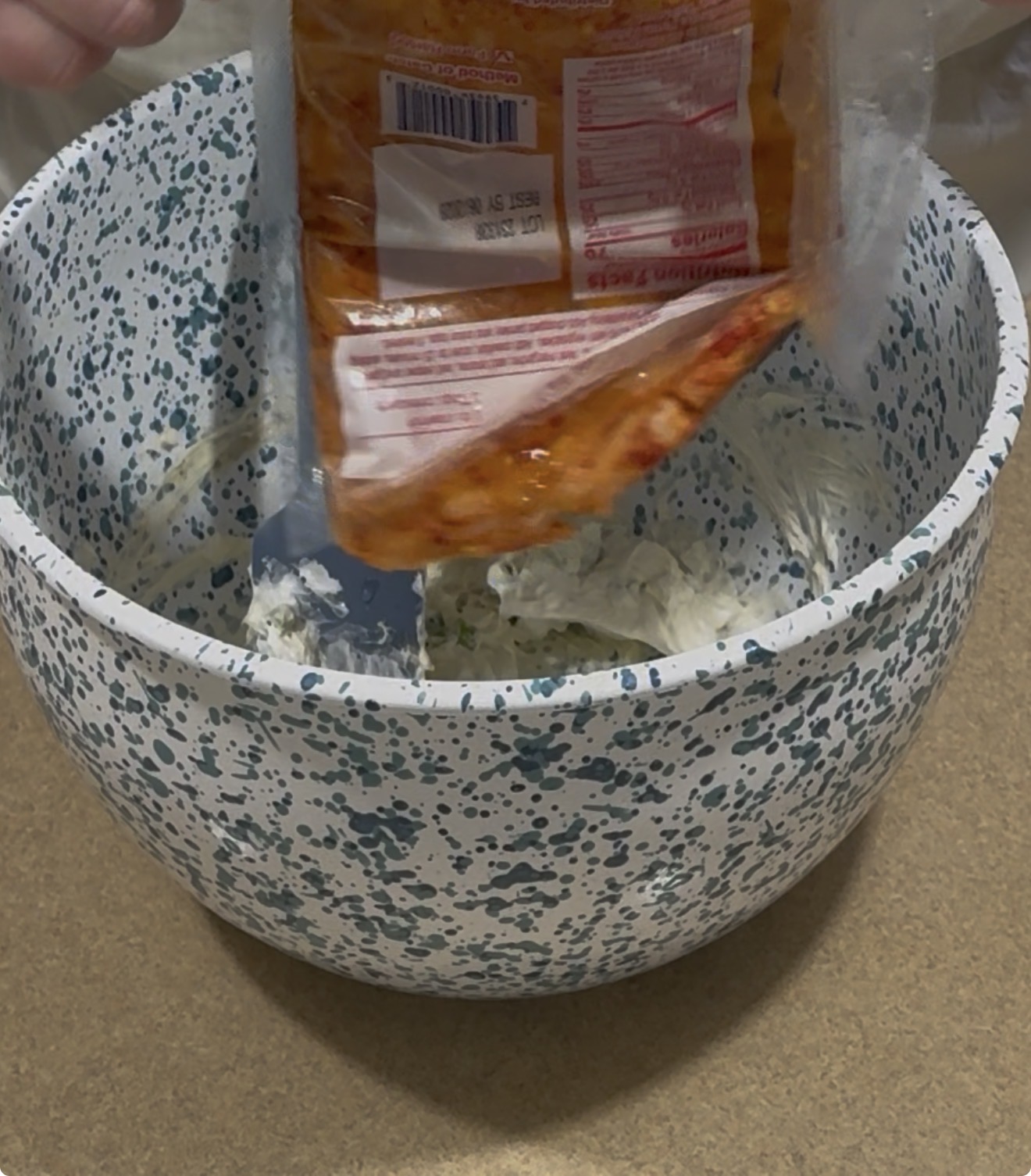 A hand squeezes the contents of a plastic packet with an orange sauce into a speckled mixing bowl containing a creamy mixture. A spatula rests inside the bowl.