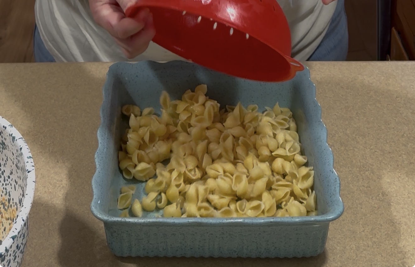 A person pours cooked pasta shells from a red strainer into a blue square baking dish on a kitchen counter.