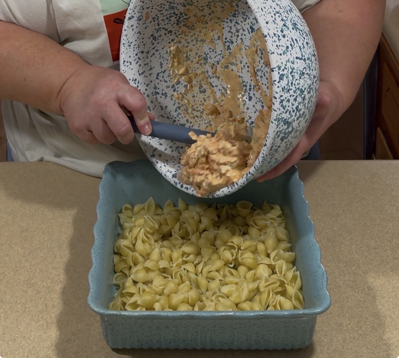 A person pours a creamy mixture from a speckled bowl onto cooked shell pasta in a blue square baking dish on a kitchen counter.