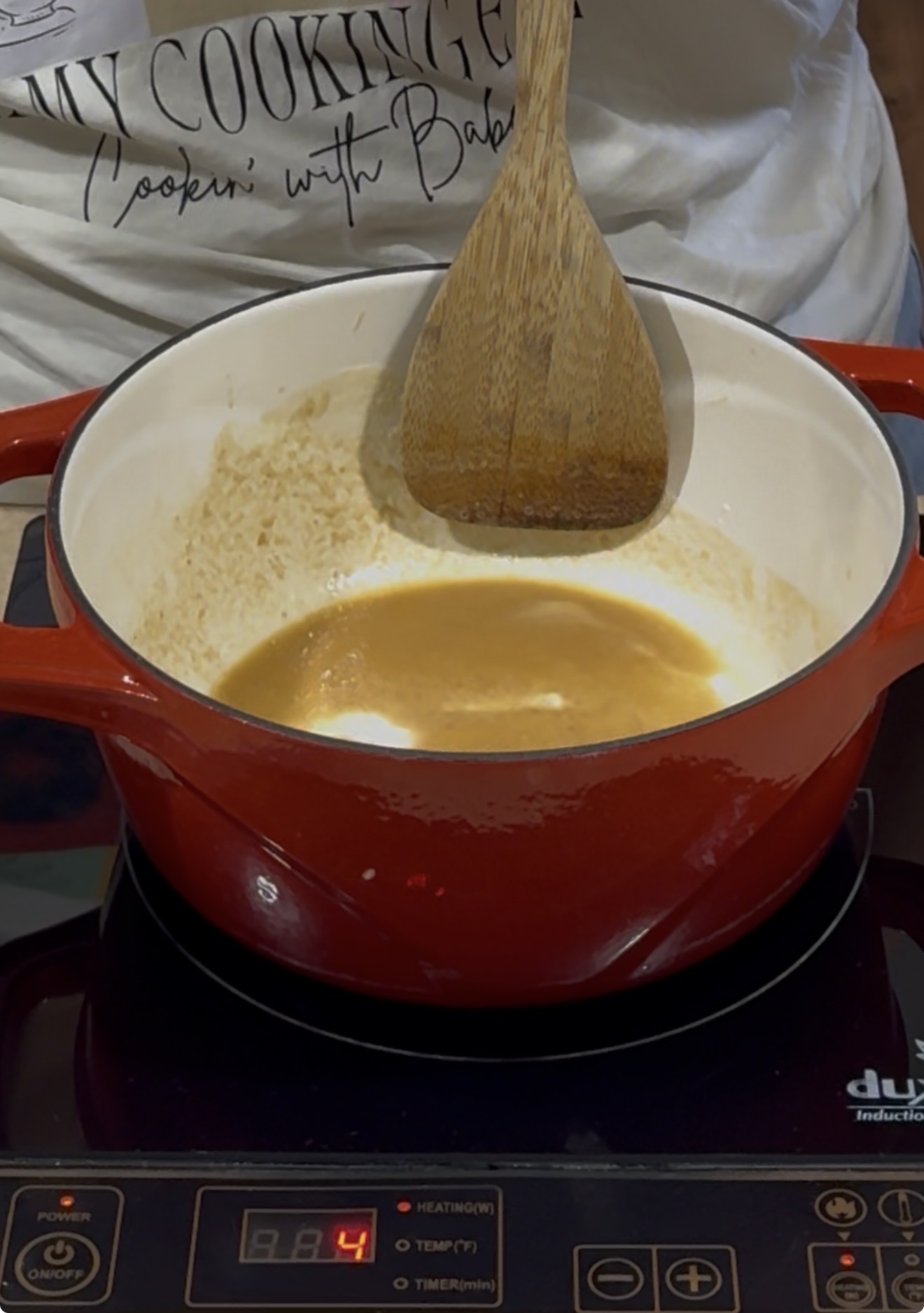 A person stirs a creamy brown sauce in a red pot with a wooden spoon on an induction cooktop. The pot is partially full, and the cooktop display shows the heat setting at 4.