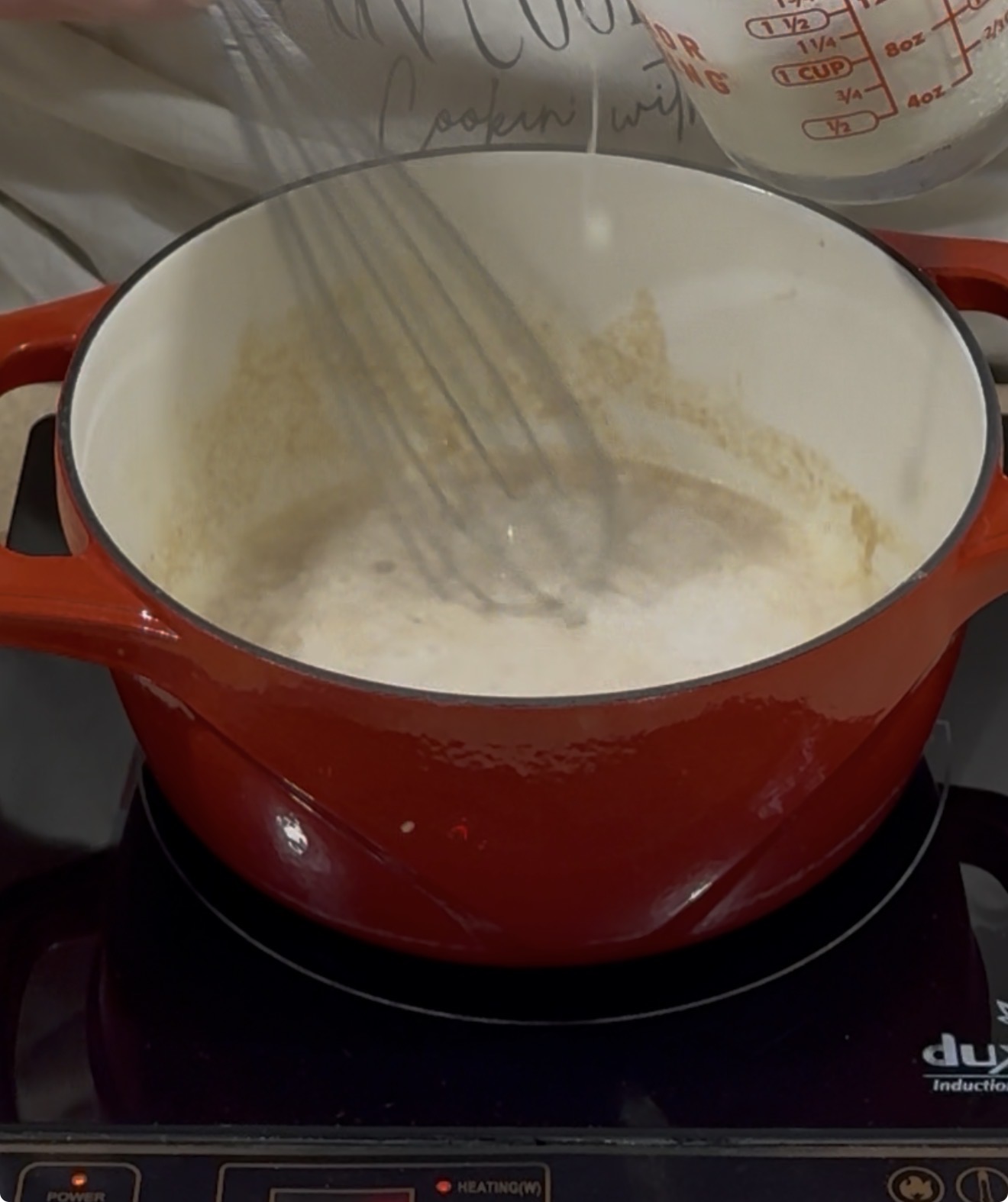 A person is whisking a creamy mixture in a red Dutch oven on a stovetop, while pouring liquid from a measuring cup into the pot.