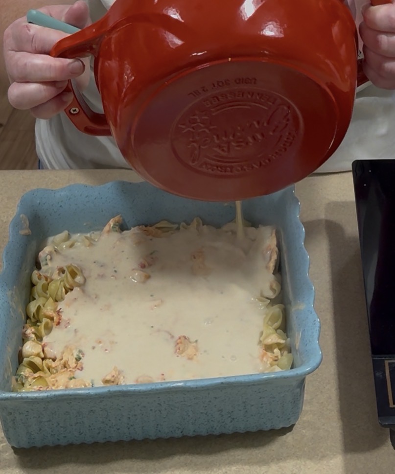 A person pours a creamy white sauce from a red mixing bowl over a pasta casserole in a blue baking dish on a kitchen counter.