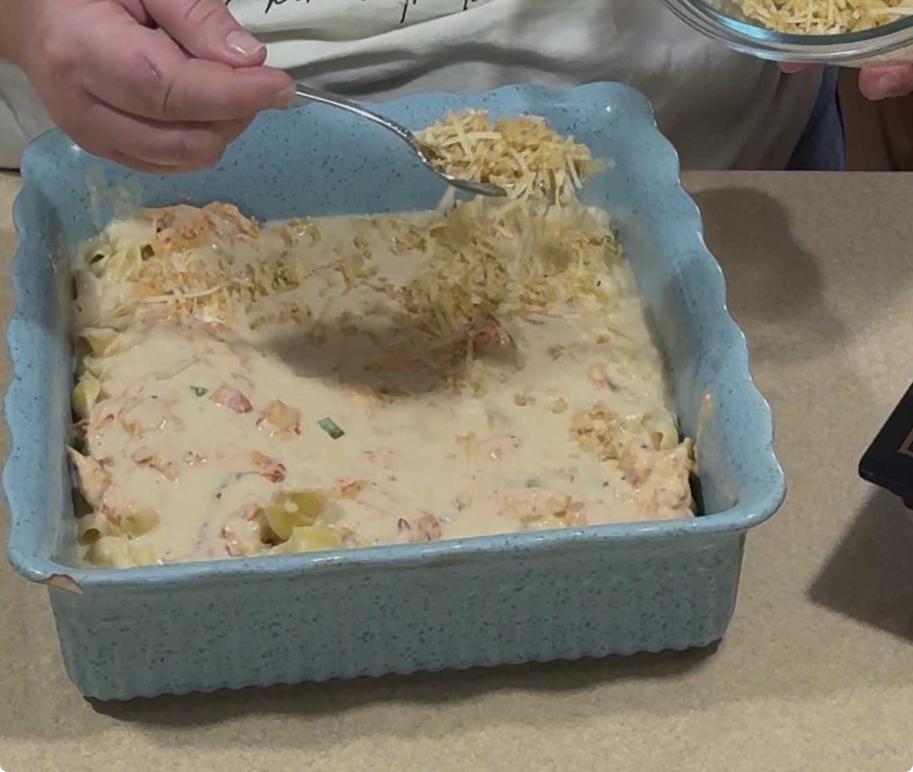 A person sprinkles shredded cheese from a spoon onto a creamy casserole in a blue square baking dish on a kitchen counter.