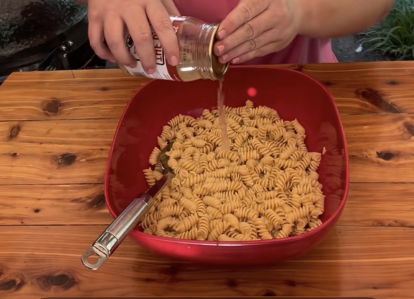 A person pours liquid from a jar onto a large bowl of cooked rotini pasta on a wooden table, with a fork resting in the bowl.