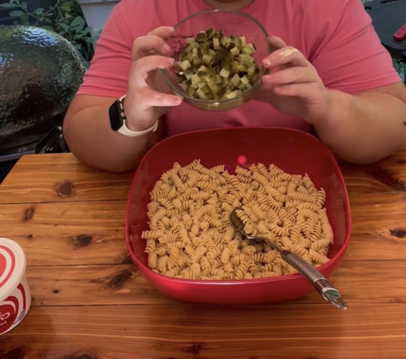 A person in a pink shirt holds a bowl of chopped pickles over a large red container filled with cooked rotini pasta on a wooden table. A container of sour cream sits nearby.