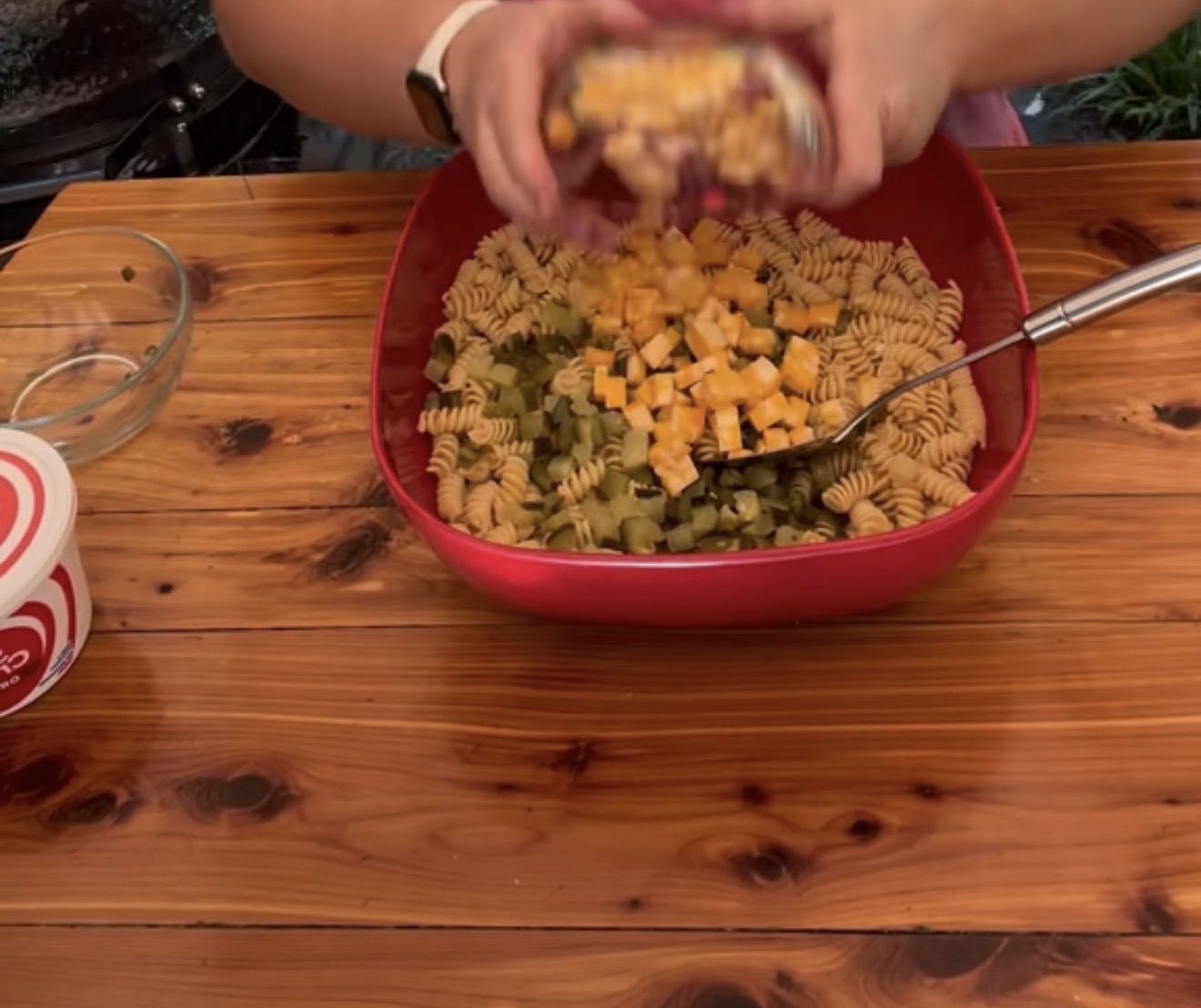 A person adds cubed cheese to a red bowl filled with rotini pasta and chopped vegetables on a wooden table. A fork, a glass bowl, and a container of sour cream are nearby.