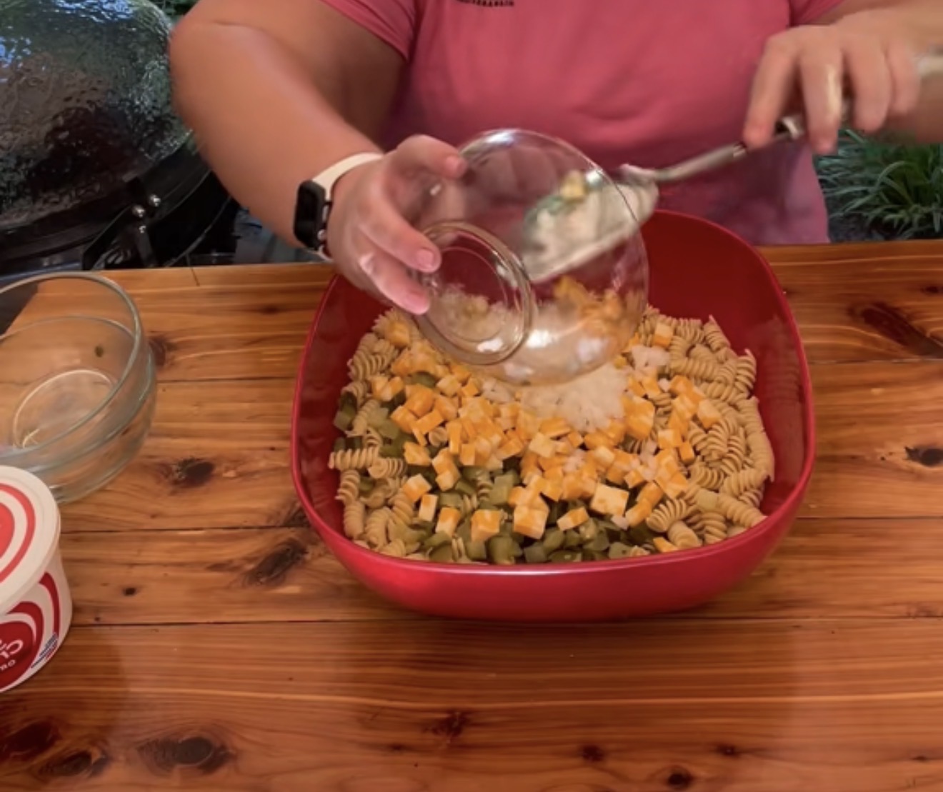 A person in a pink shirt pours chopped onions into a red bowl containing rotini pasta, cubed cheese, and pickles on a wooden table, preparing a dish outdoors.