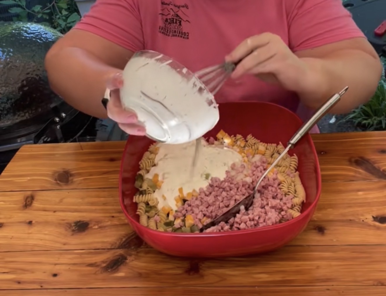 A person in a pink shirt pours creamy dressing from a bowl into a large red bowl filled with cooked rotini pasta, diced ham, cheese cubes, and vegetables on a wooden table.