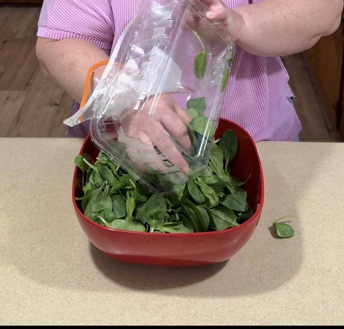 A person in a pink striped shirt pours fresh spinach leaves from a plastic container into a red bowl on a beige kitchen counter.