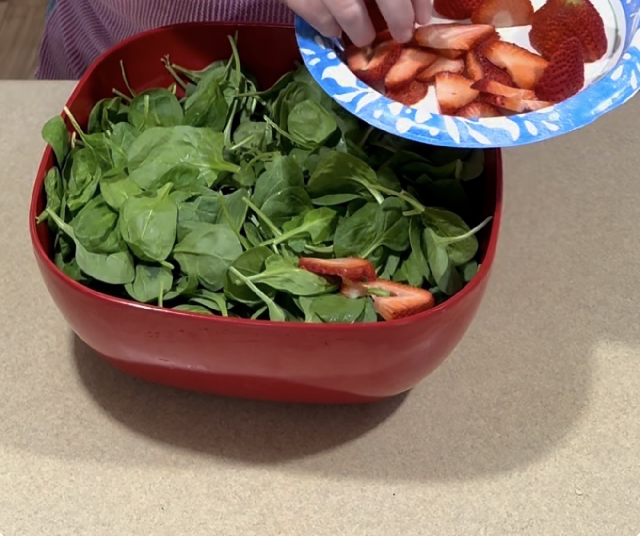 A hand adds sliced strawberries from a paper plate into a large red bowl filled with fresh spinach leaves on a countertop.